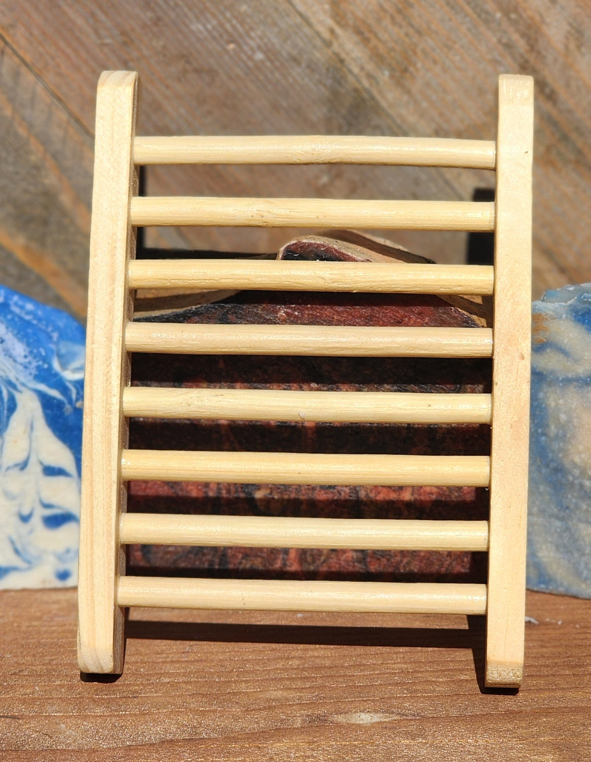 Light wooden soap dish with slatted rails, standing upright in front of a bar of soap, with blue and white marbled soap bars on either side
