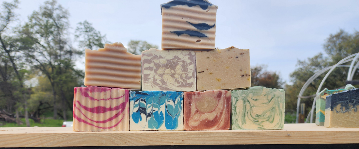 A pyramid arrangement of artisanal handmade soaps in various marbled and swirled patterns, displayed on a wooden surface with a blurred outdoor background of trees and sky.