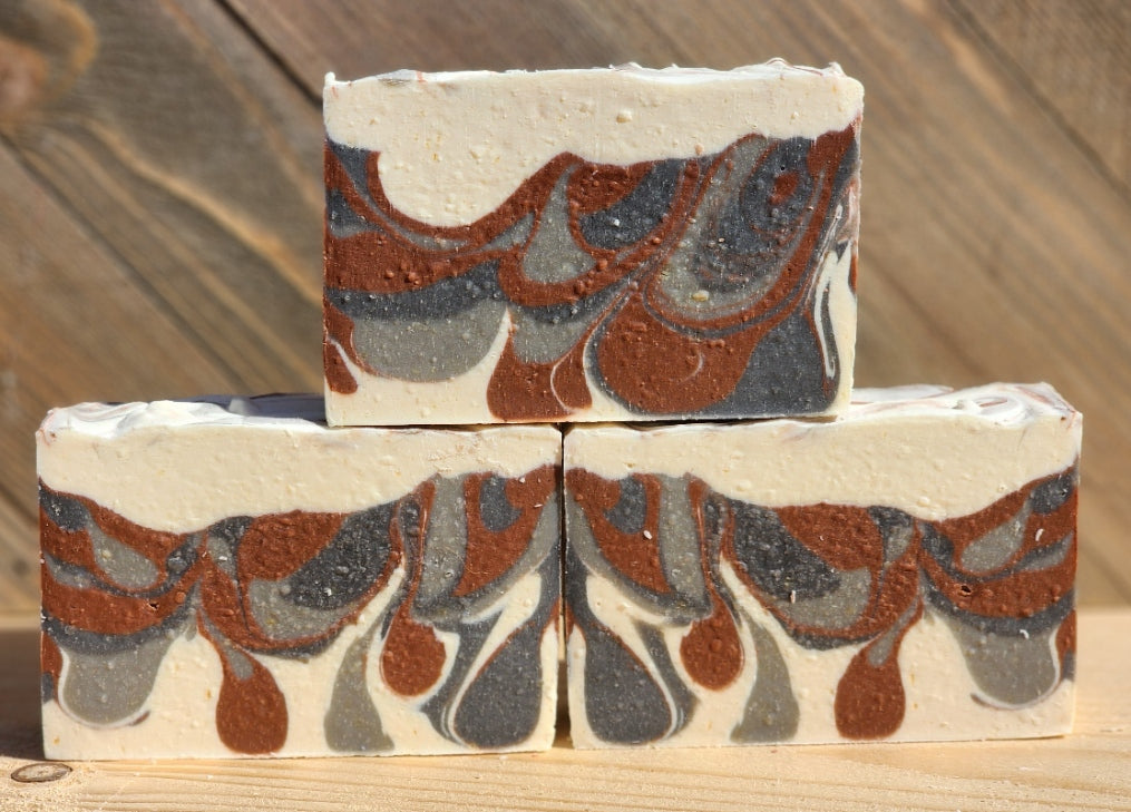 Three rectangular bars of handmade soap stacked in a pyramid arrangement on a wooden surface, featuring a marbled pattern of cream, terracotta brown, and dark gray.