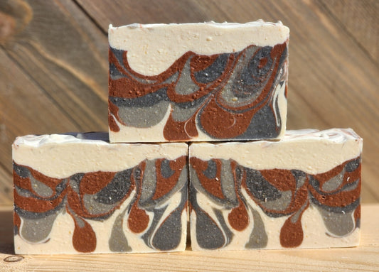 Three rectangular bars of handmade soap stacked in a pyramid arrangement on a wooden surface, featuring a marbled pattern of cream, terracotta brown, and dark gray.