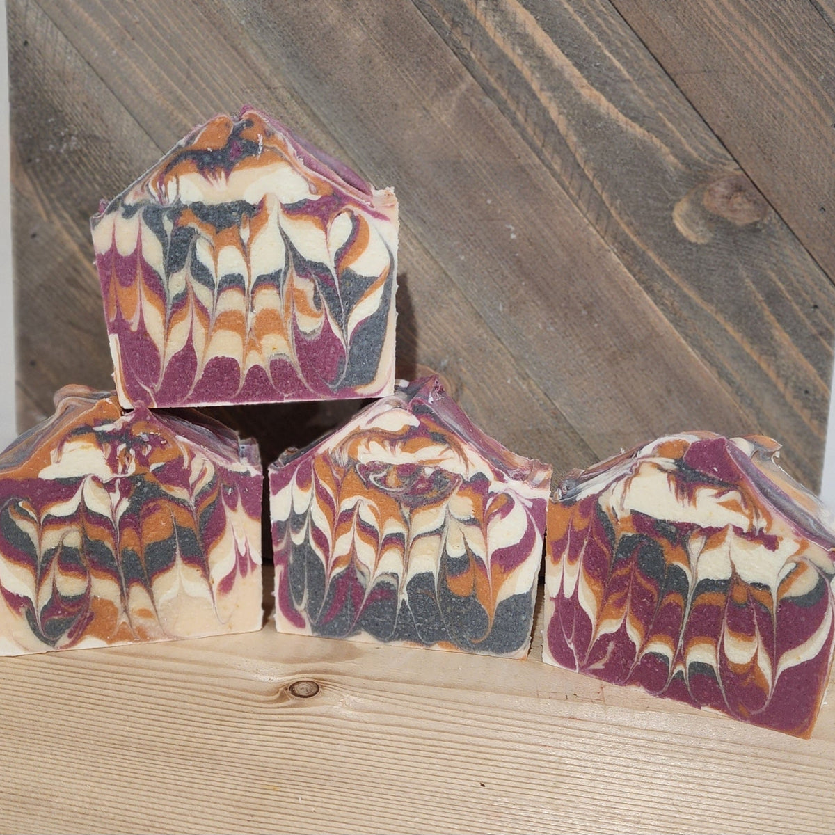 Four handmade soap bars stacked on a wooden surface, with feathered swirls of orange, purple, cream, and dark gray.
