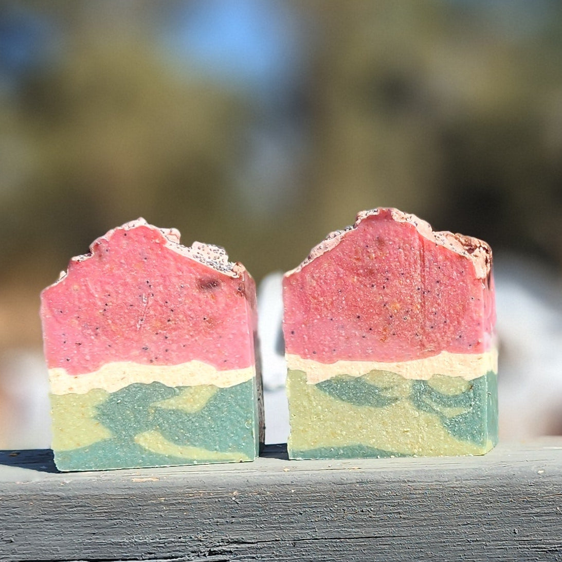 Two layered soap bars with pink, green, and white colors on a wooden surface with a blurred natural background.