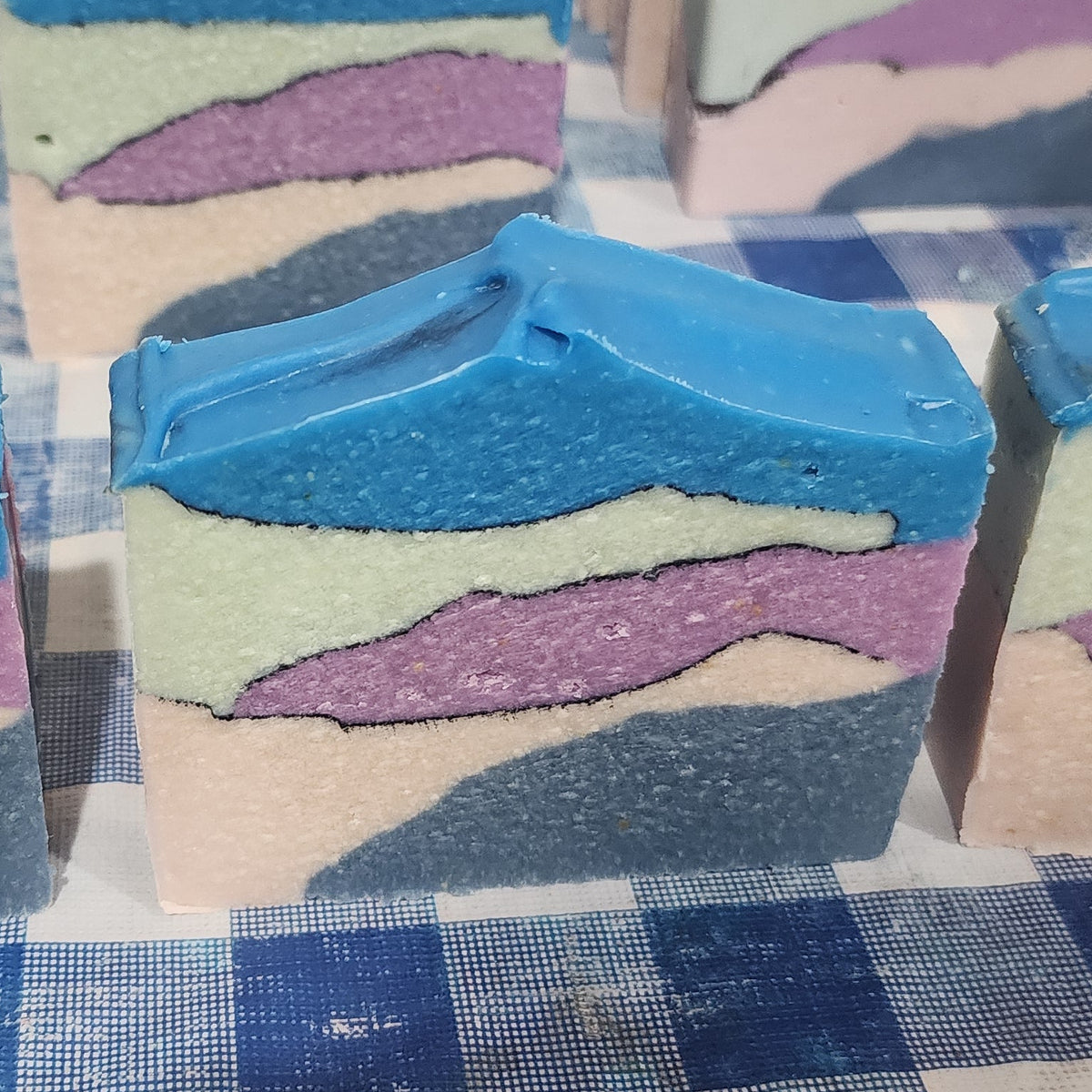 Tipsy Lane goat milk soap: layered bars in green, purple, and pink with a bright blue top, on a checkered towel.