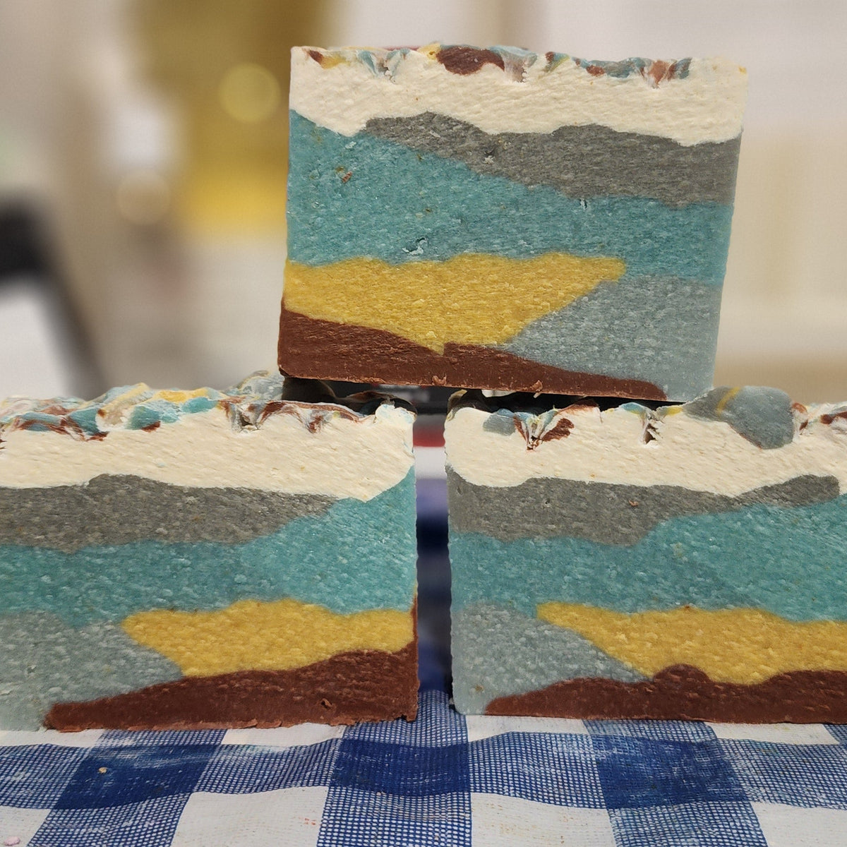 Three layered soap bars with a mountain landscape design on a checkered tablecloth.