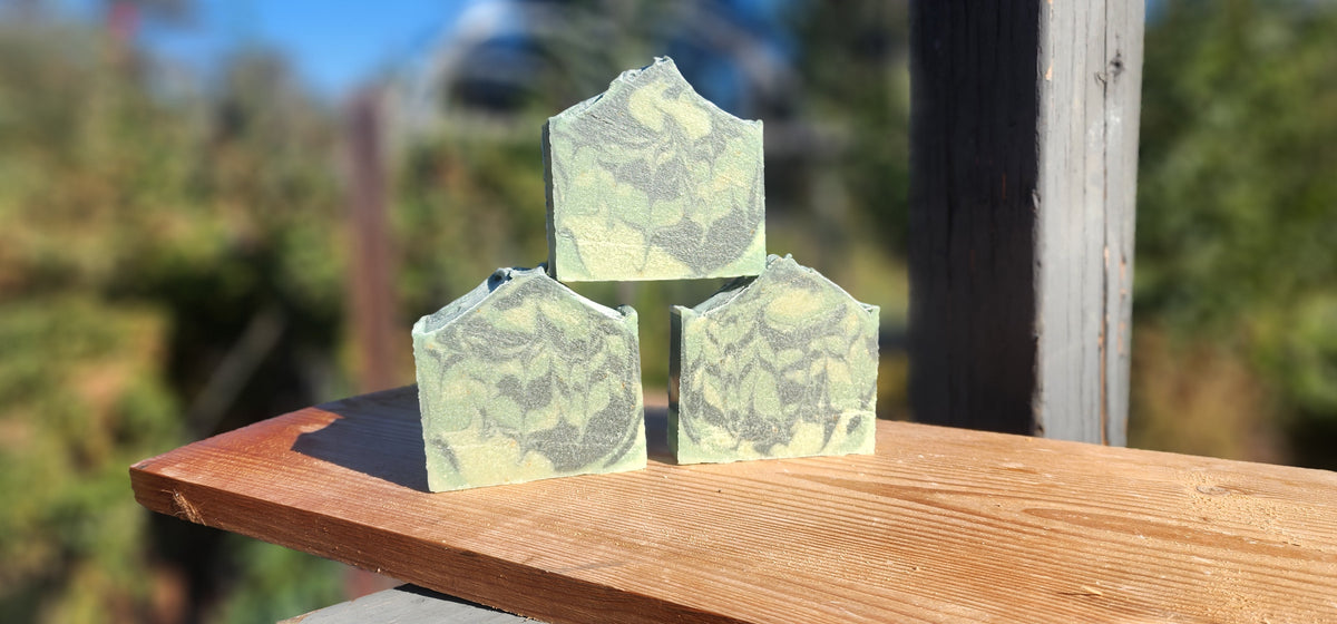 Three bars of green soap stacked on a wooden board outdoors, with blurred greenery in the background.