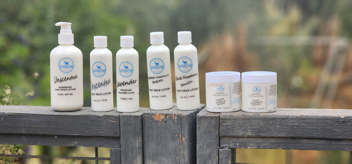 Six white personal care products from Shattuck Farm Goat Milk Soap N Stuff lined up on a weathered wooden railing, with a soft, blurred outdoor garden background.