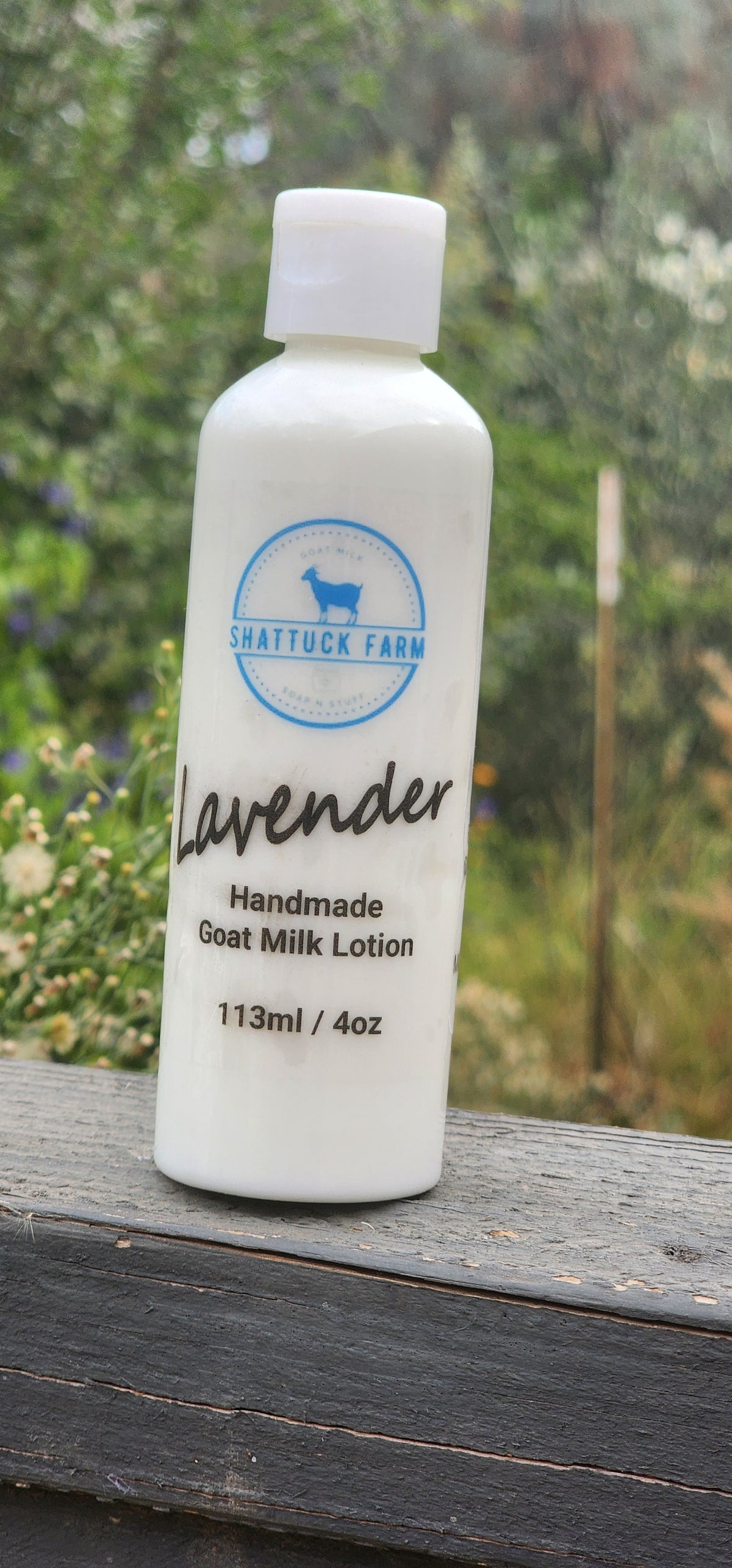 White 4 oz squeeze bottle of Shattuck Farm Lavender Handmade Goat Milk Lotion on a weathered wooden railing with a blurred garden background