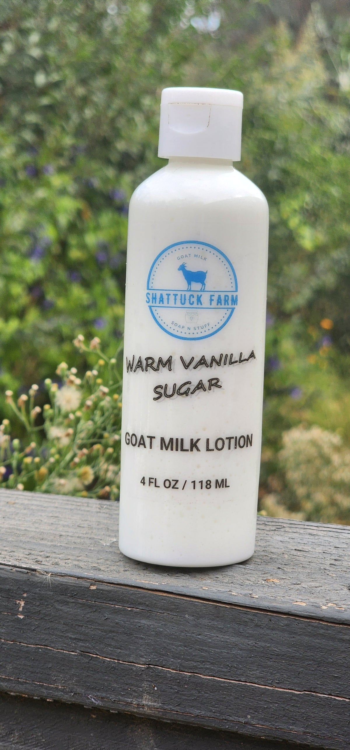 A white bottle of Shattuck Farm Warm Vanilla Sugar Goat Milk Lotion sitting on a dark wooden surface with a blurred green garden background.