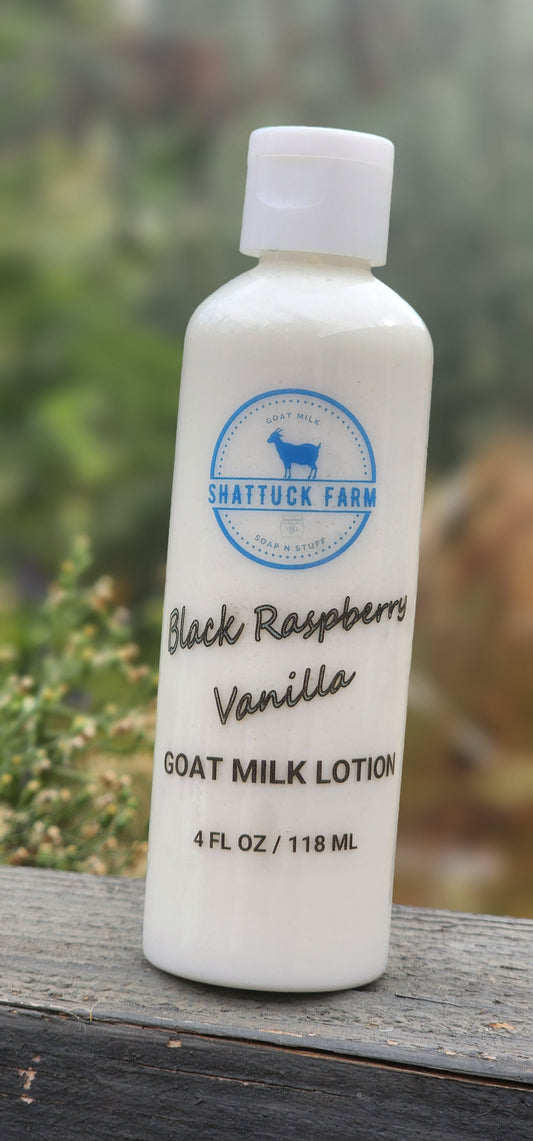 A white bottle of Shattuck Farm Black Raspberry Vanilla Goat Milk Lotion, 4 fl oz, standing on a weathered wooden surface with a blurred outdoor garden background.