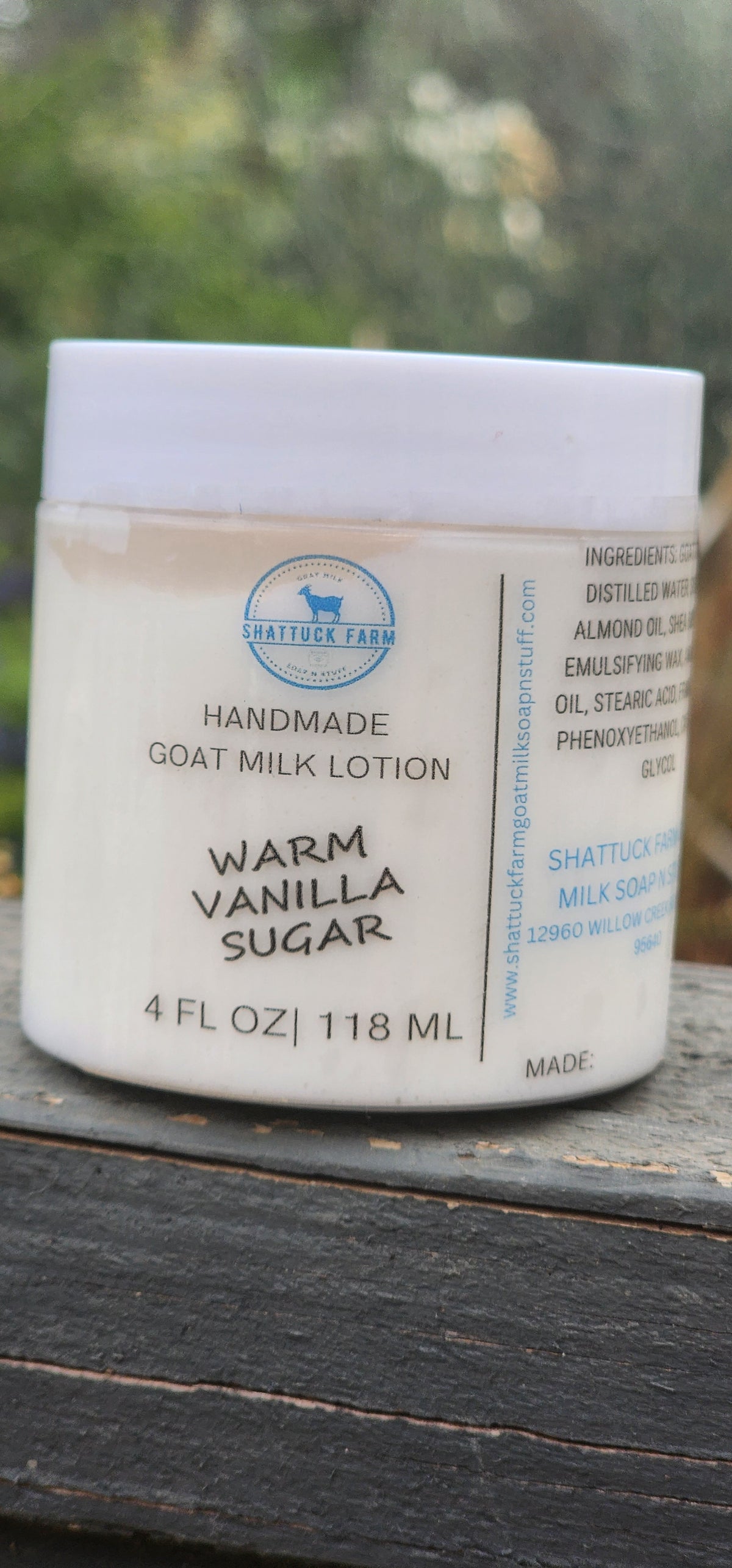 A jar of Shattuck Farm Handmade Goat Milk Lotion in Warm Vanilla Sugar scent, resting on a dark wooden surface with a blurred green outdoor background.