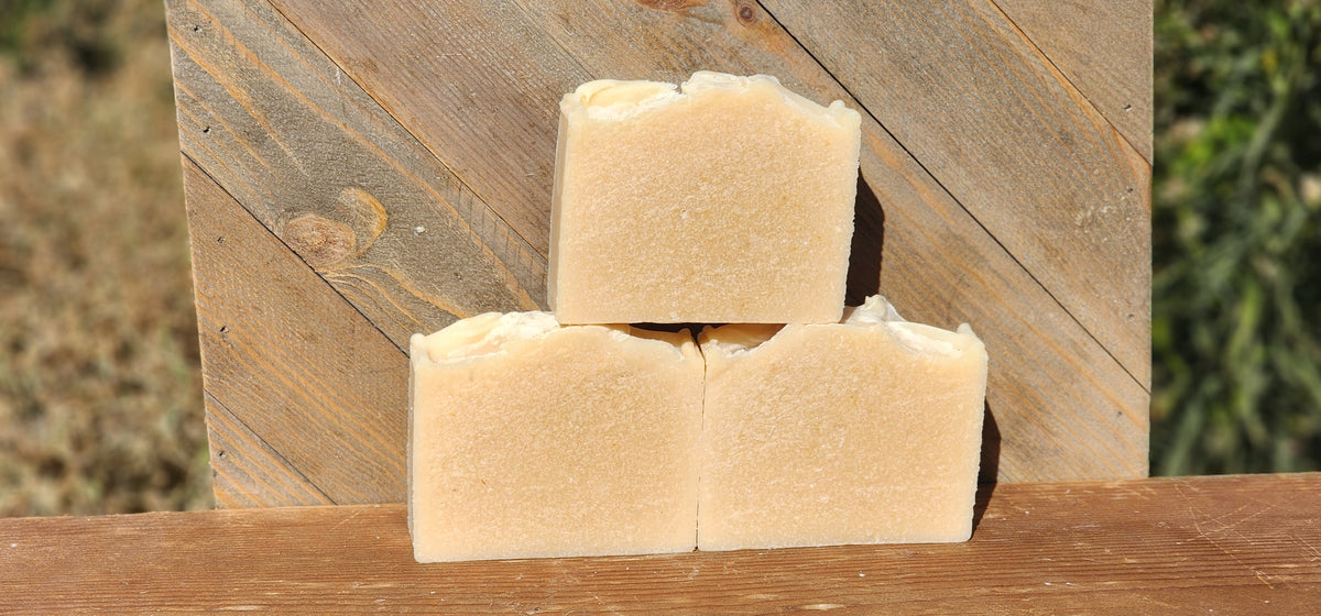 Unscented goat milk soap: three rectangular bars of pale beige soap stacked in a pyramid arrangement on a wooden surface, with a rustic wooden plank background and blurred greenery behind it.