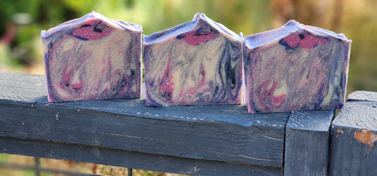 Black Raspberry Vanilla goat milk soap: three rectangular bars of marbled purple, pink, and cream soap resting on a weathered dark blue wooden fence outdoors.