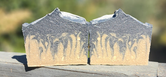 Two rectangular bars of handmade soap with a marbled gray and beige pattern, displayed side-by-side on a weathered wooden railing with a blurred outdoor background.