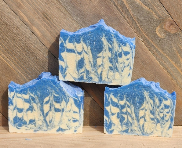 Dutch Delph goat milk soap: three handmade marbled soap bars with blue and cream swirls, arranged on a light wood surface against a darker wood plank background.