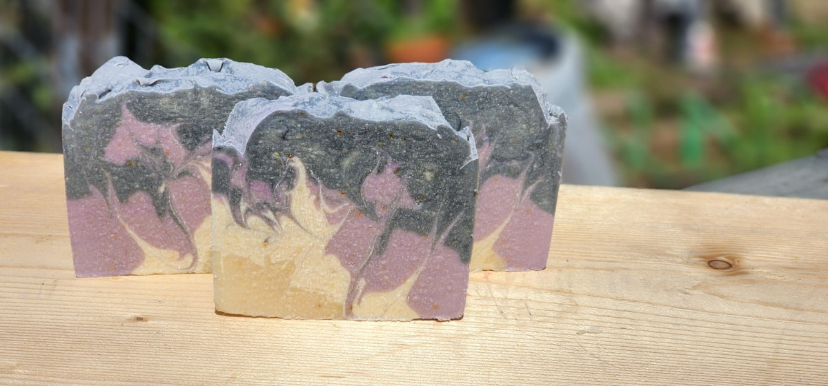 Purple Iris goat milk soap: three handmade soap bars with a swirled pattern of black, dusty purple, and beige, resting on a light wooden surface with a blurred outdoor background.