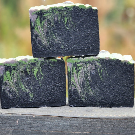 Spooky Night (Champagne Toast) goat milk soap: three black bars with green swirls and a frosted top, stacked in a pyramid on a weathered wooden surface with a blurred garden background.