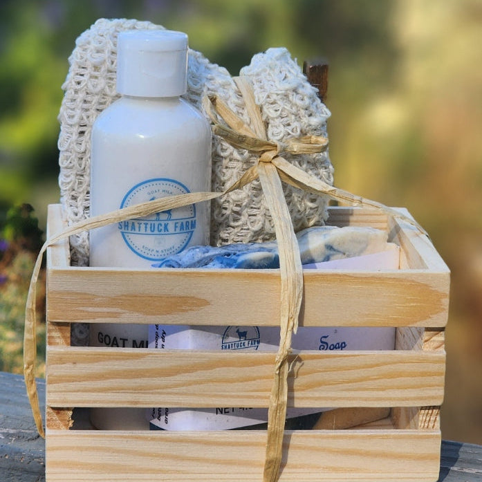 A wooden gift basket filled with bath and body items, including a bar of goat milk soap, white bottle of goat milk lotion, a textured cream towel or sponge, and wrapped accessories, tied with a tan ribbon, set on a wooden railing with a blurred outdoor background.