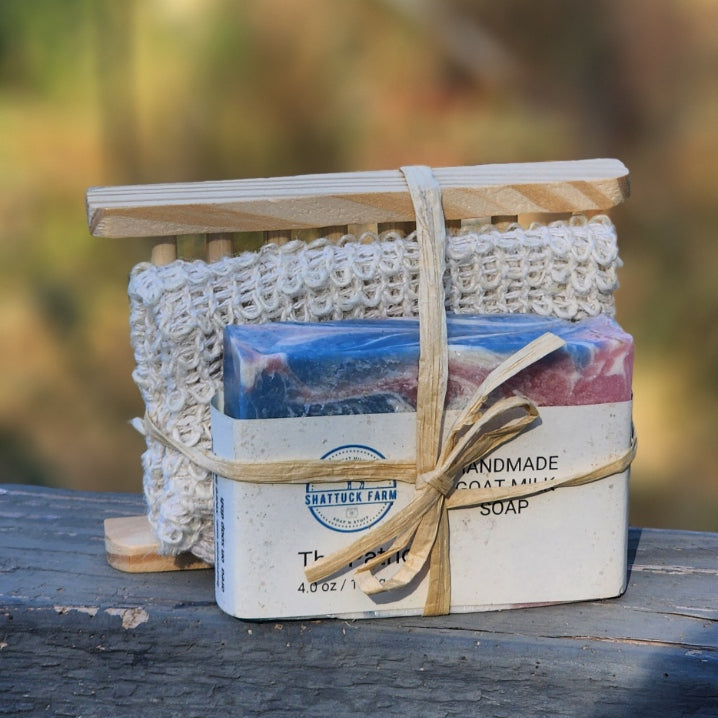 A packaged handmade soap gift set wrapped in a rustic tan ribbon, resting on a weathered wooden surface with a soft, blurred outdoor background.