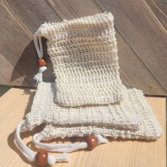 Two woven beige sisal soap bags with wooden beads on their drawstrings, lying on a light wooden surface against a gray wooden background.
