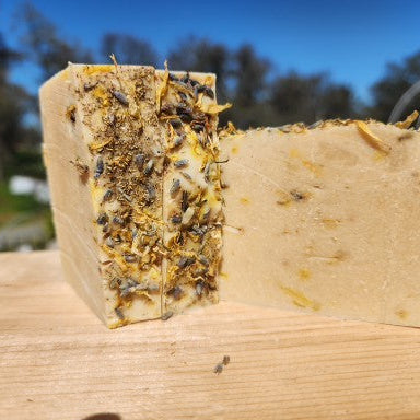 Summer Wildflowers goat milk soap: two bars on a wooden surface outdoors, one topped with dried flower petals and one plain cream bar.